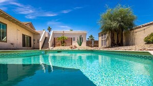 Una piscina con agua azul cristalina, rodeada de edificios con paredes de estuco y techos de tejas, y una palmera al fondo.
