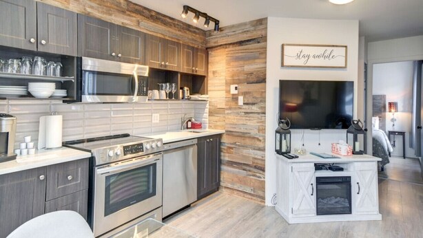 A modern kitchen with stainless steel appliances, wooden wall accents, and a mounted TV. - Chouette Tremblant Ski In/Out Pedestrian Village (Mont Tremblant)