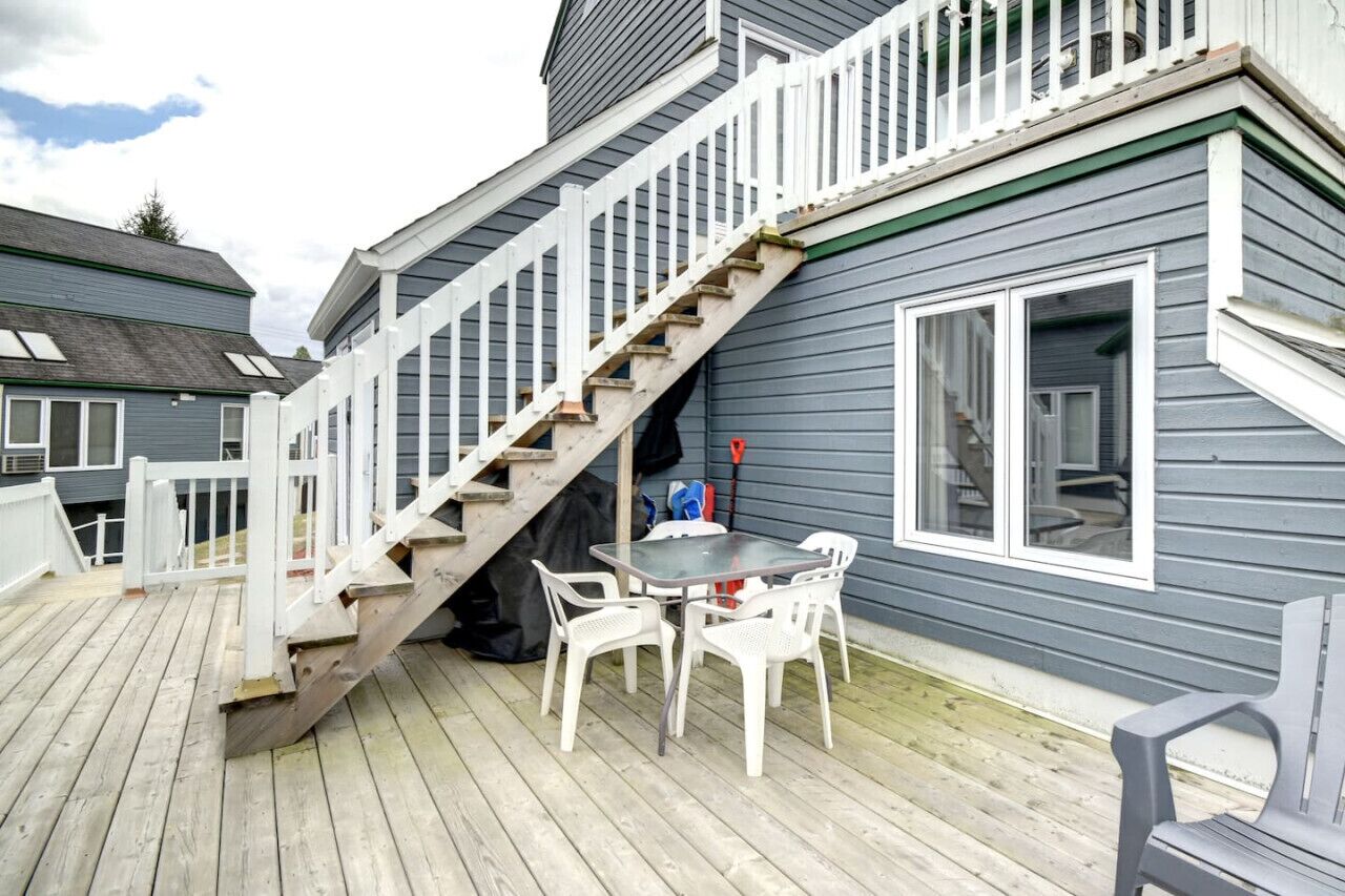 A wooden deck with a table and chairs, a staircase leading to a second floor, and a window with blinds.