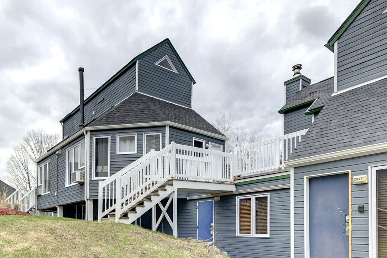 A two-story residential building with a prominent gable roof, a front porch with white railings, and a blue door.