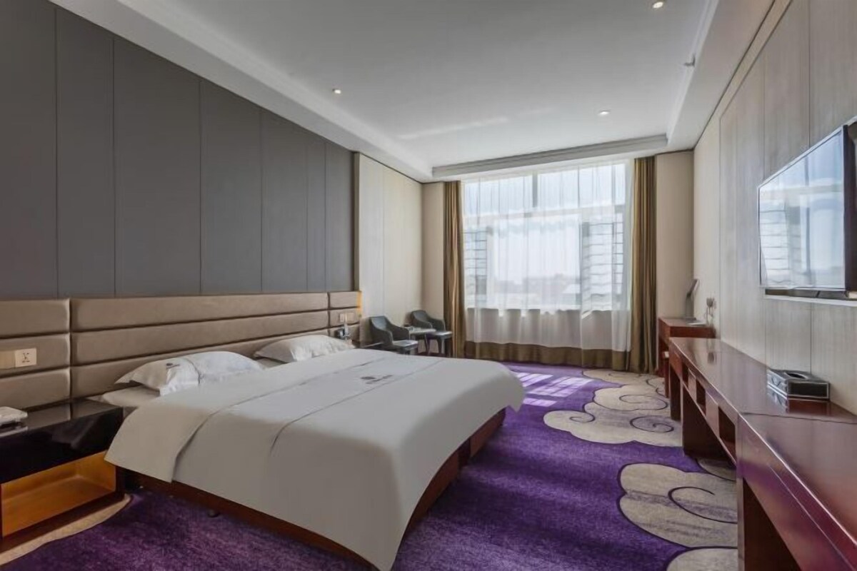 A hotel room with a large bed, a desk with a chair, a TV, and a window with curtains.