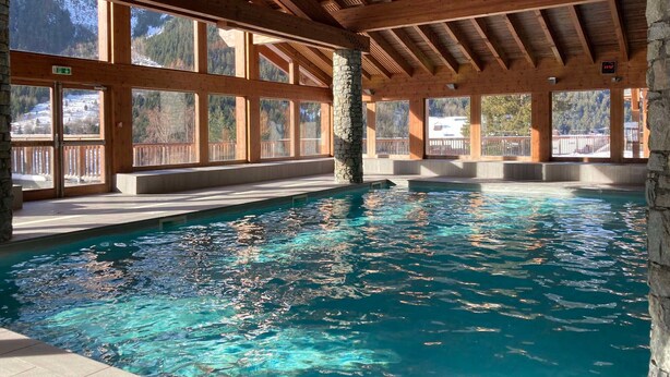 An indoor swimming pool with wooden beams and large windows offering views of a snowy mountain landscape. - Beautiful apartment with balcony and covered parking (Pralognan-la-Vanoise)
