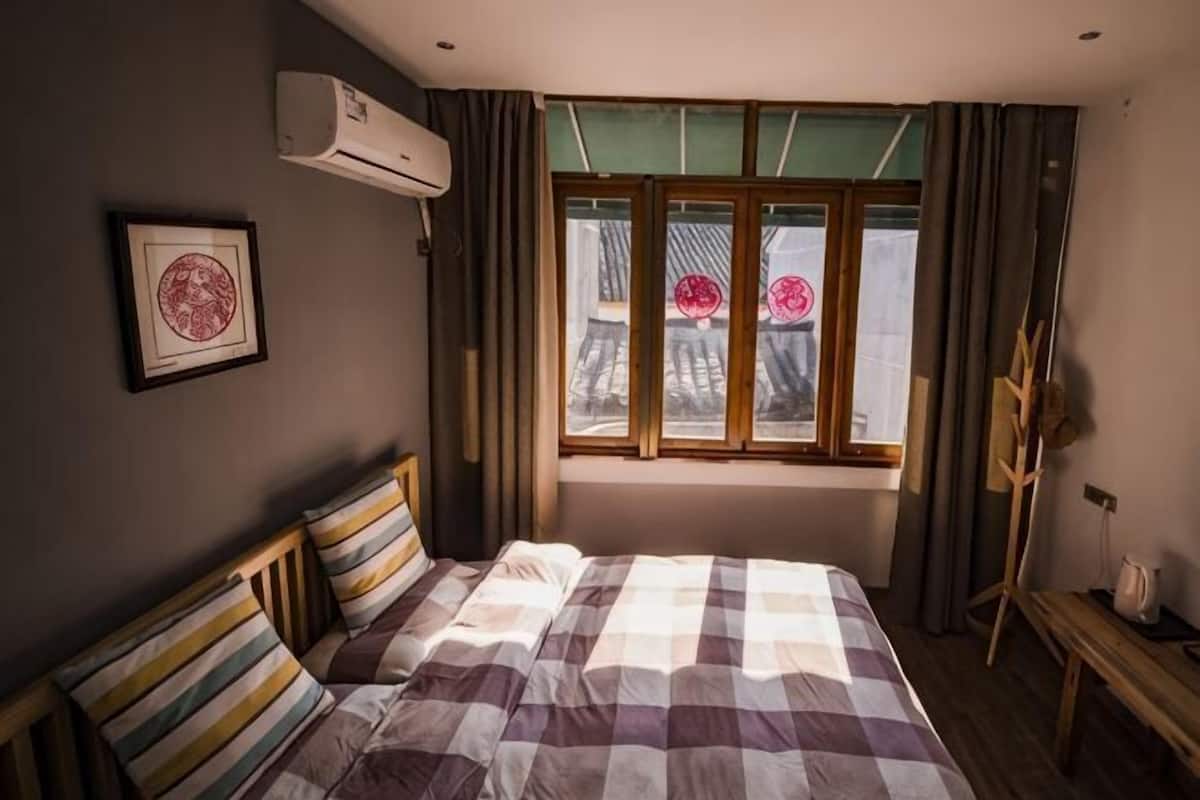 Quarto com cama, janela com cortinas, ar condicionado fixado na parede e um quadro emoldurado.