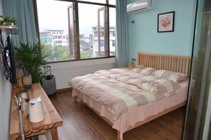 Un dormitorio con cama, un escritorio de madera, una planta en maceta y una ventana con vistas a edificios.