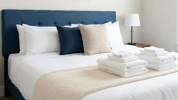 A neatly made bed with a blue headboard, white and beige pillows, and a stack of white towels.