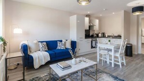 A modern living room with a blue sofa, white dining table, and a kitchen in the background.