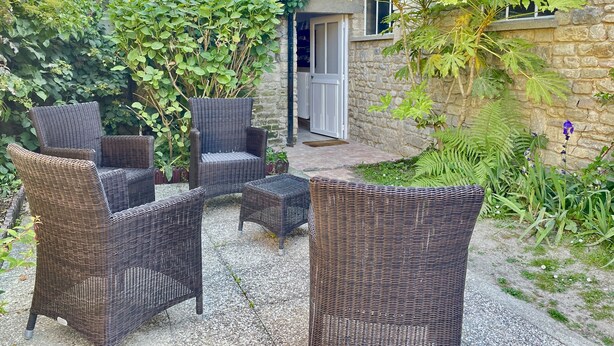 A stone building with a garden patio featuring wicker furniture and lush greenery. - L'orangerie des Ursulines (Bayeux)