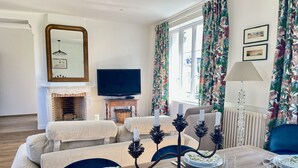 A living room with a fireplace, a television, floral curtains, and a dining area set for a meal. - L'orangerie des Ursulines (Bayeux)