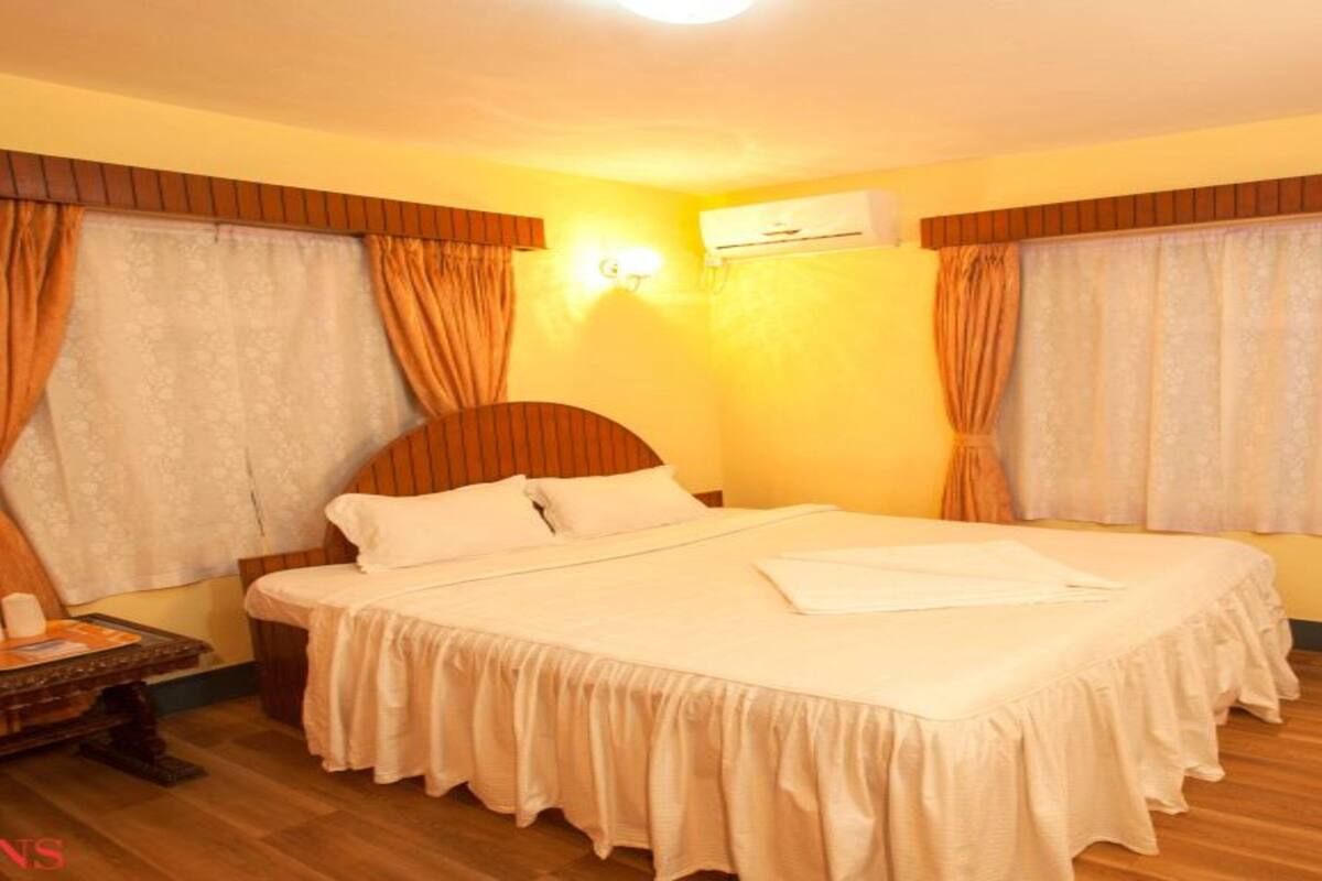 A hotel room with a large bed, two windows with curtains, an air conditioning unit, and a bedside table with a lamp.