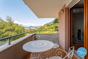 A balcony with a round table and chairs, a glass door, and a view of greenery and buildings. - Apartment T2 – Clos Charmeil – Lake (Veyrier-du-Lac)