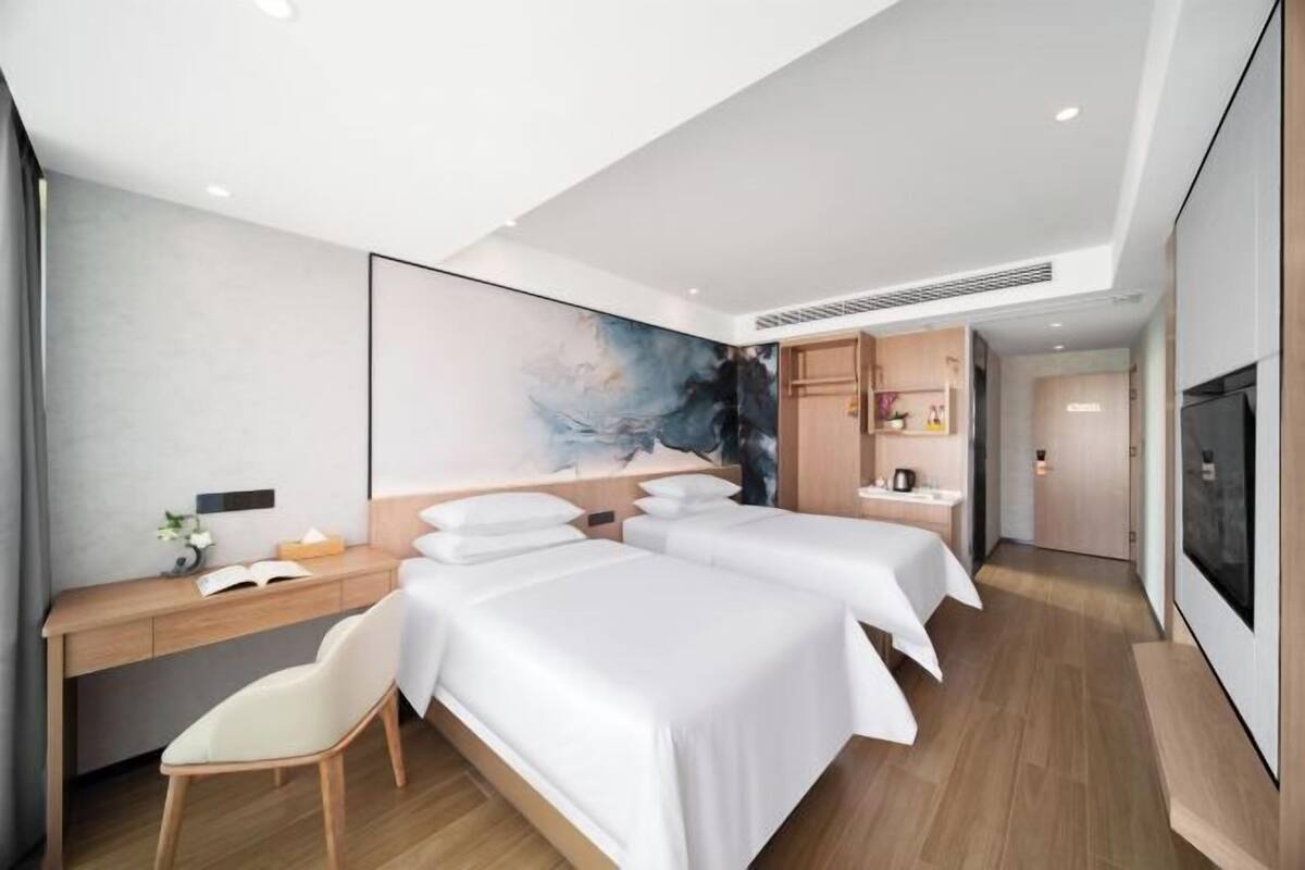 A modern hotel room with two beds, a desk, a chair, a large artwork on the wall, and a television.