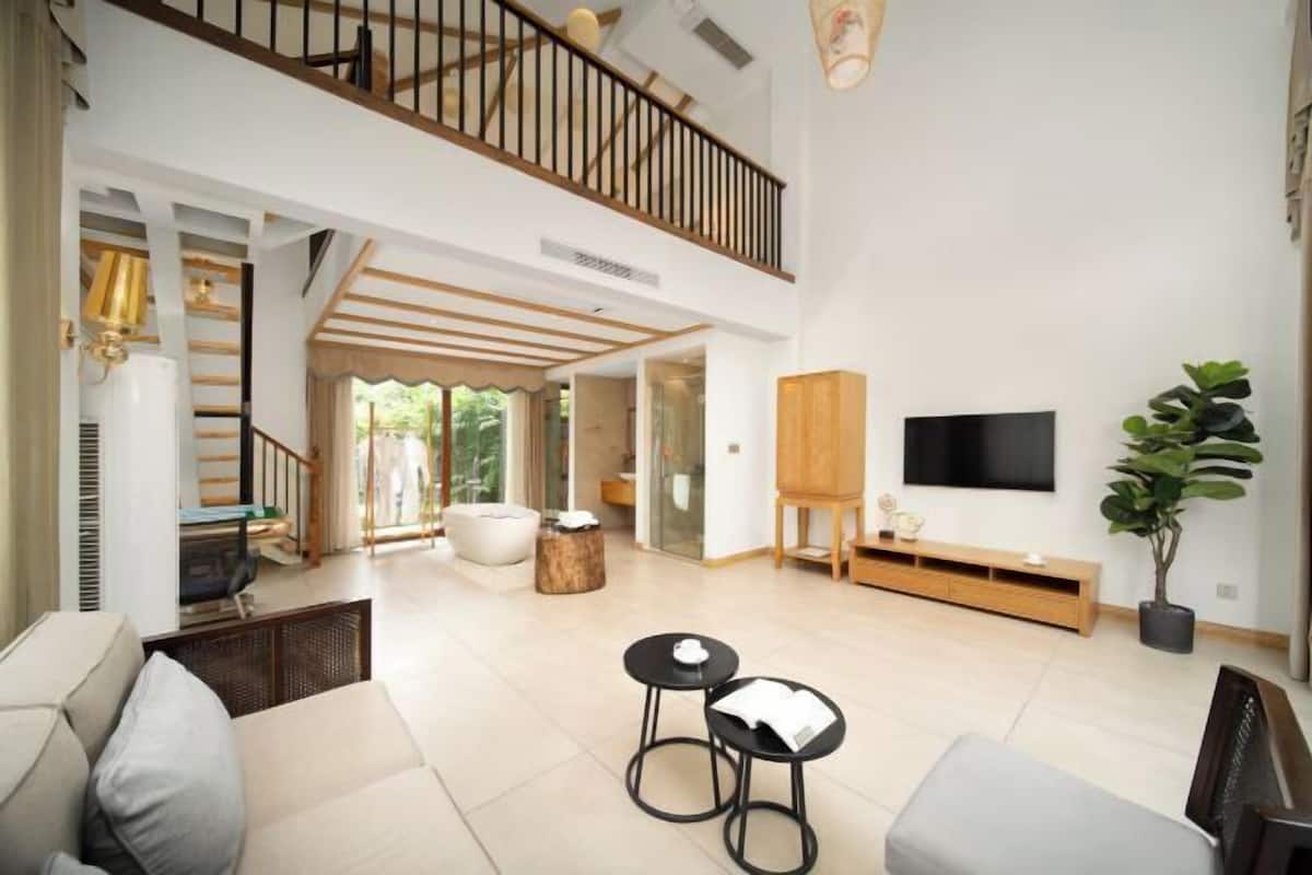 A spacious living room with a high ceiling, a staircase, a flat-screen TV, a wooden cabinet, and a potted plant.