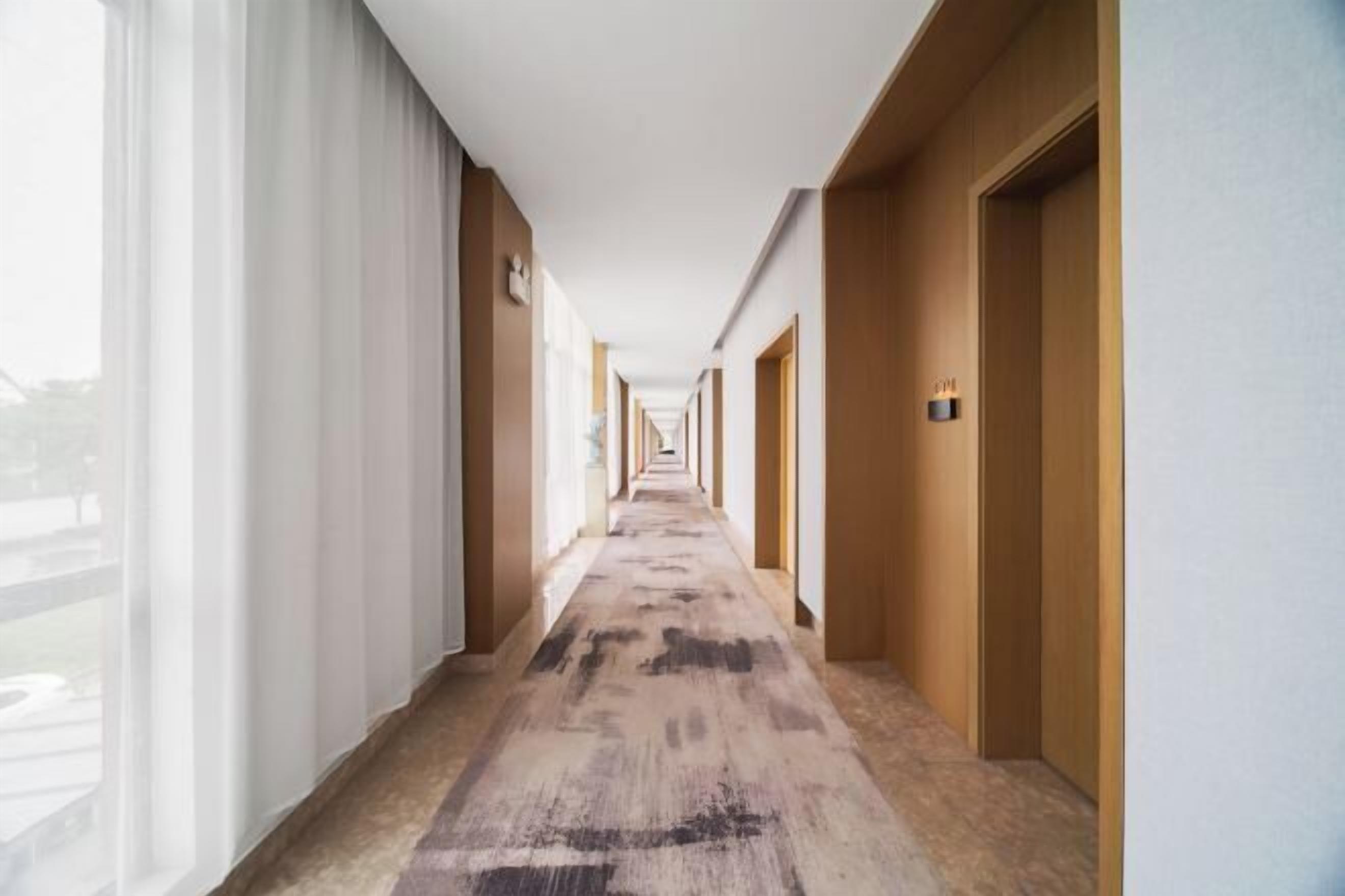 Um corredor de hotel com piso coberto por carpete, portas de madeira e uma janela à esquerda.