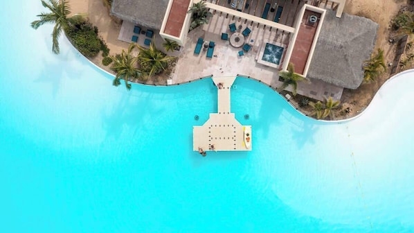 An aerial view of a resort with a central pool, a thatched-roof building, and palm trees.
