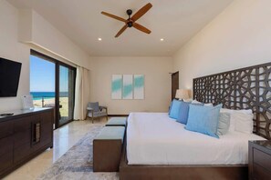 A bedroom with a large bed, a ceiling fan, a TV, and a view of the beach.