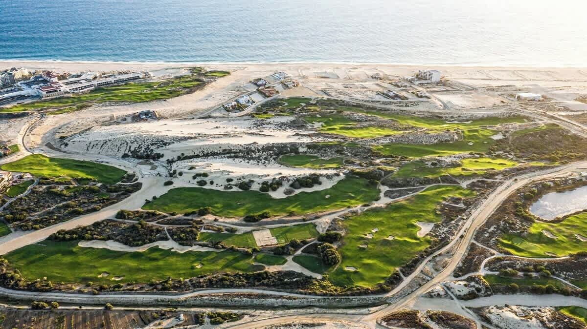 An aerial view of a coastal area with a golf course, pathways, and buildings near the beach.