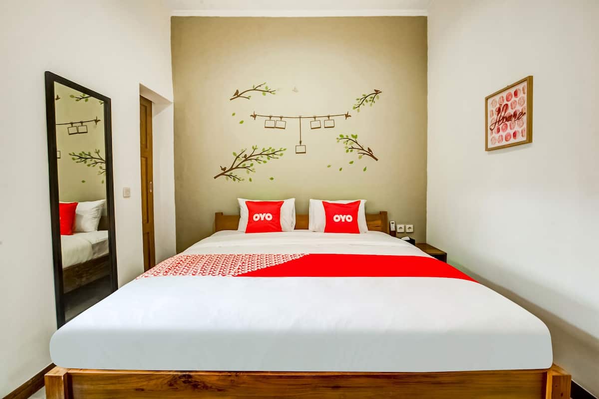 Ein Hotelzimmer mit einem großen Bett, zwei roten Kissen mit "OYO"-Branding, einer roten und weißen Tagesdecke, einem Spiegel links und einer Wand mit einer Lichterkette und einem gerahmten Bild.