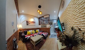 Een gezellige woonkamer met een bank, salontafel en decoratieve kussens. Een flatscreen televisie hangt aan de muur, en er staat een plant in de hoek.