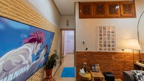 A room with a large anime-style painting on the wall, a brick wall, a wooden cabinet, a table with a lamp, and a framed photo collection. - Maison Fifi (Drâa-Tafilalet)