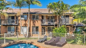 A two-story building with a swimming pool, lounge chairs, and palm trees.