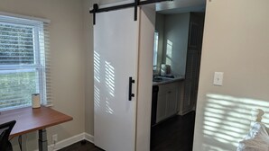 A compact living space with a sliding door leading to a kitchen, a dining area with a table and chair, and a window with blinds.