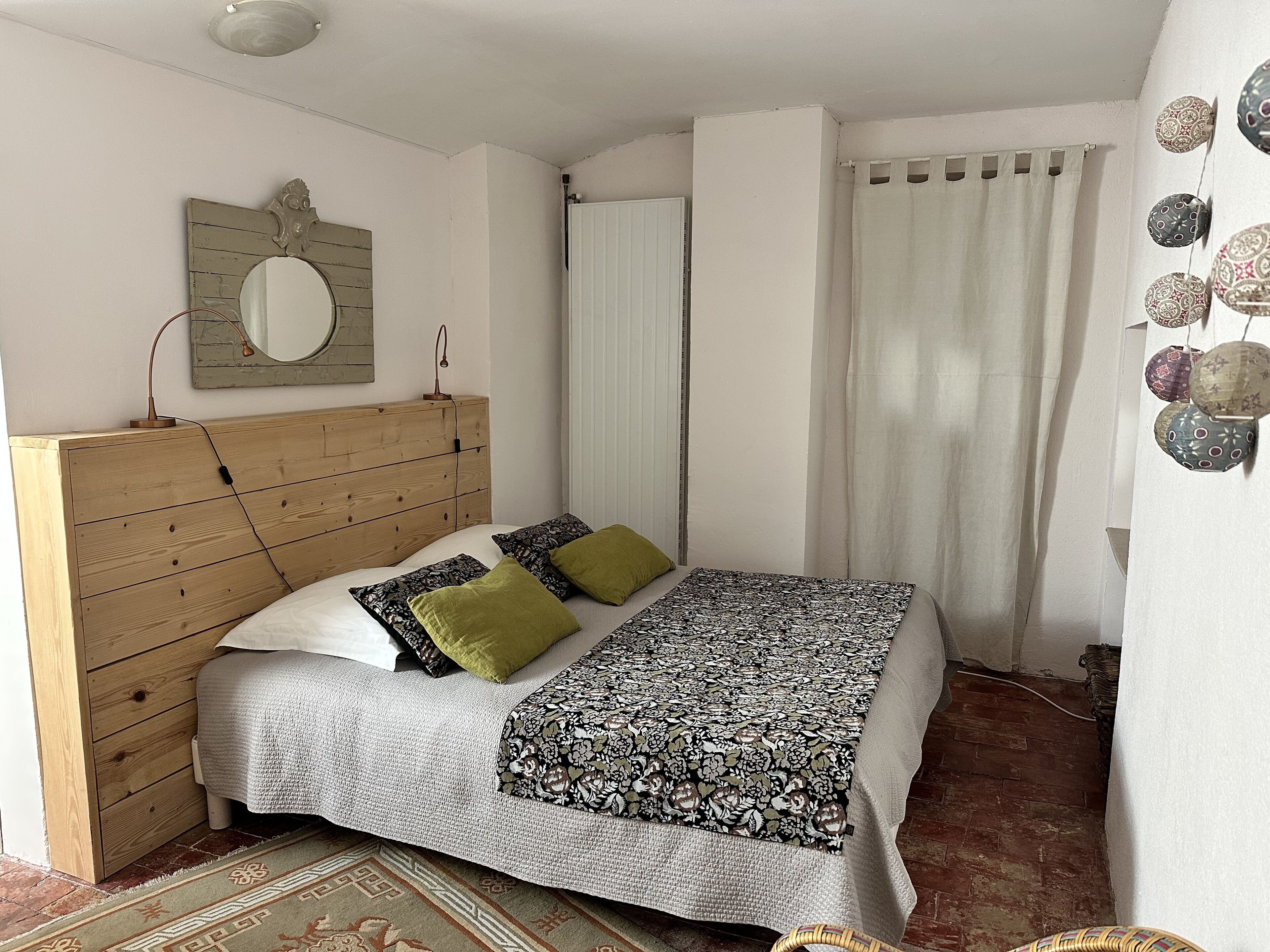 A bedroom with a wooden headboard, a bed with patterned bedding, a mirror, a radiator, and a curtain.