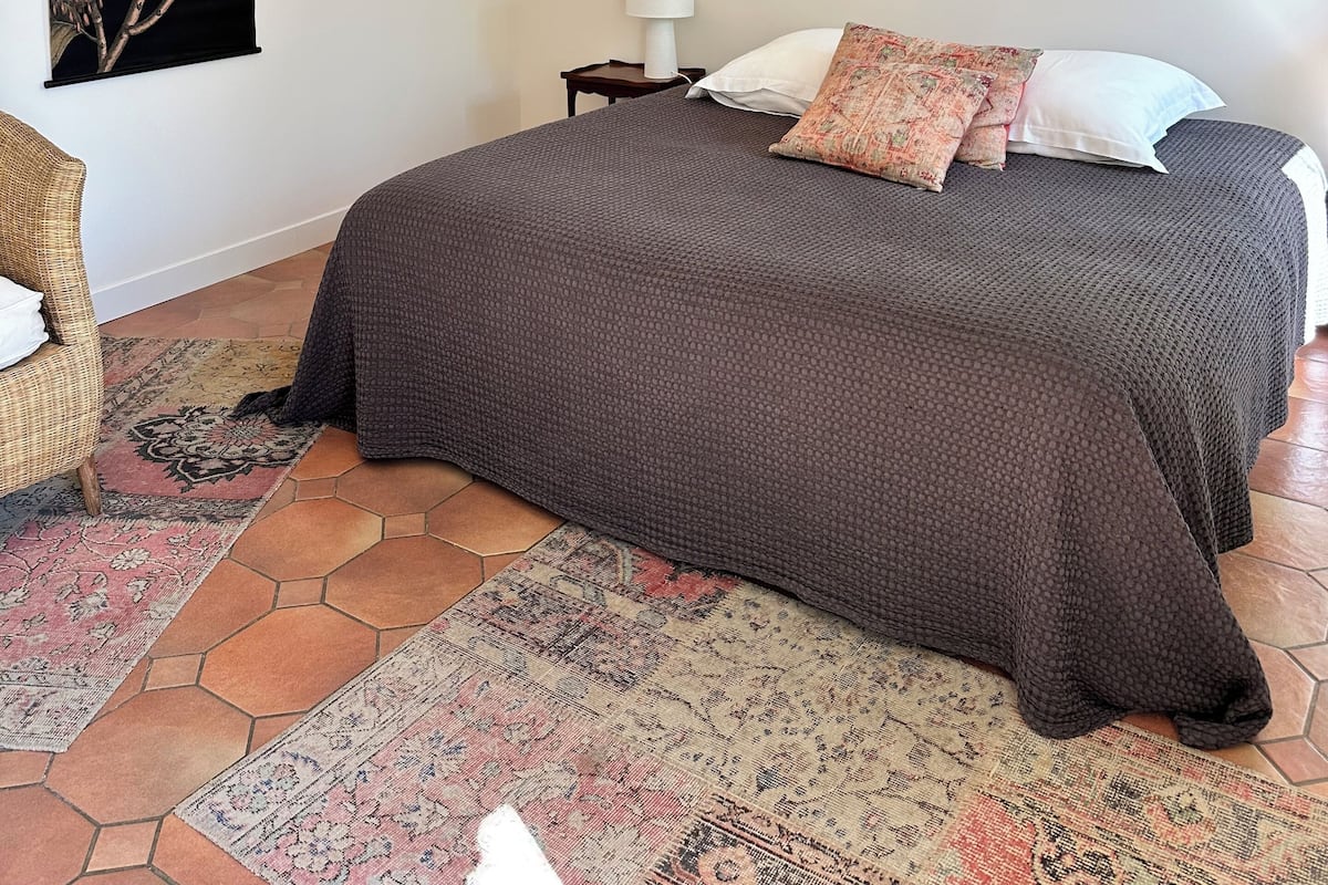 A bedroom with a large bed, a nightstand, a lamp, a chair, and a wall-mounted artwork.
