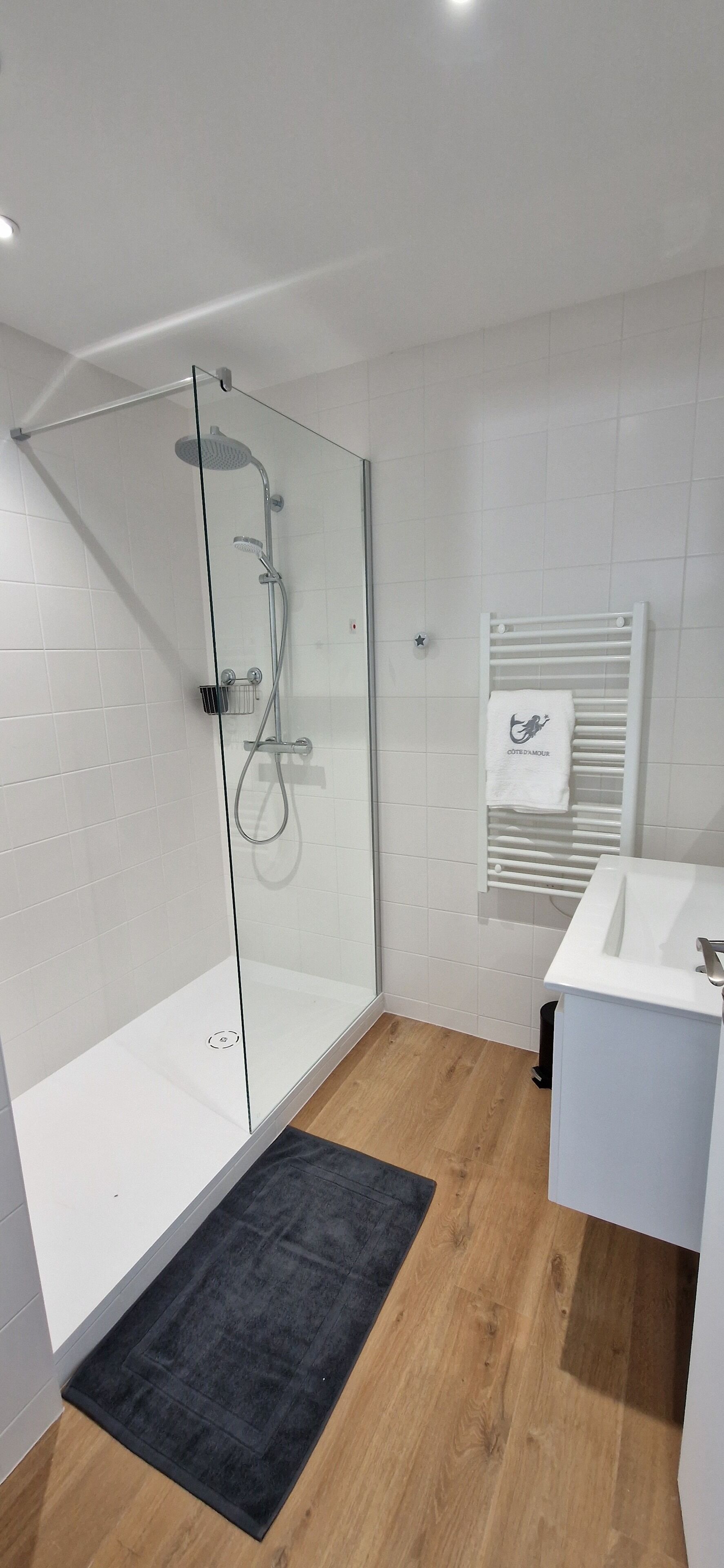 Un baño moderno con ducha envuelta en vidrio, azulejos blancos, suelo de madera y lavabo blanco.