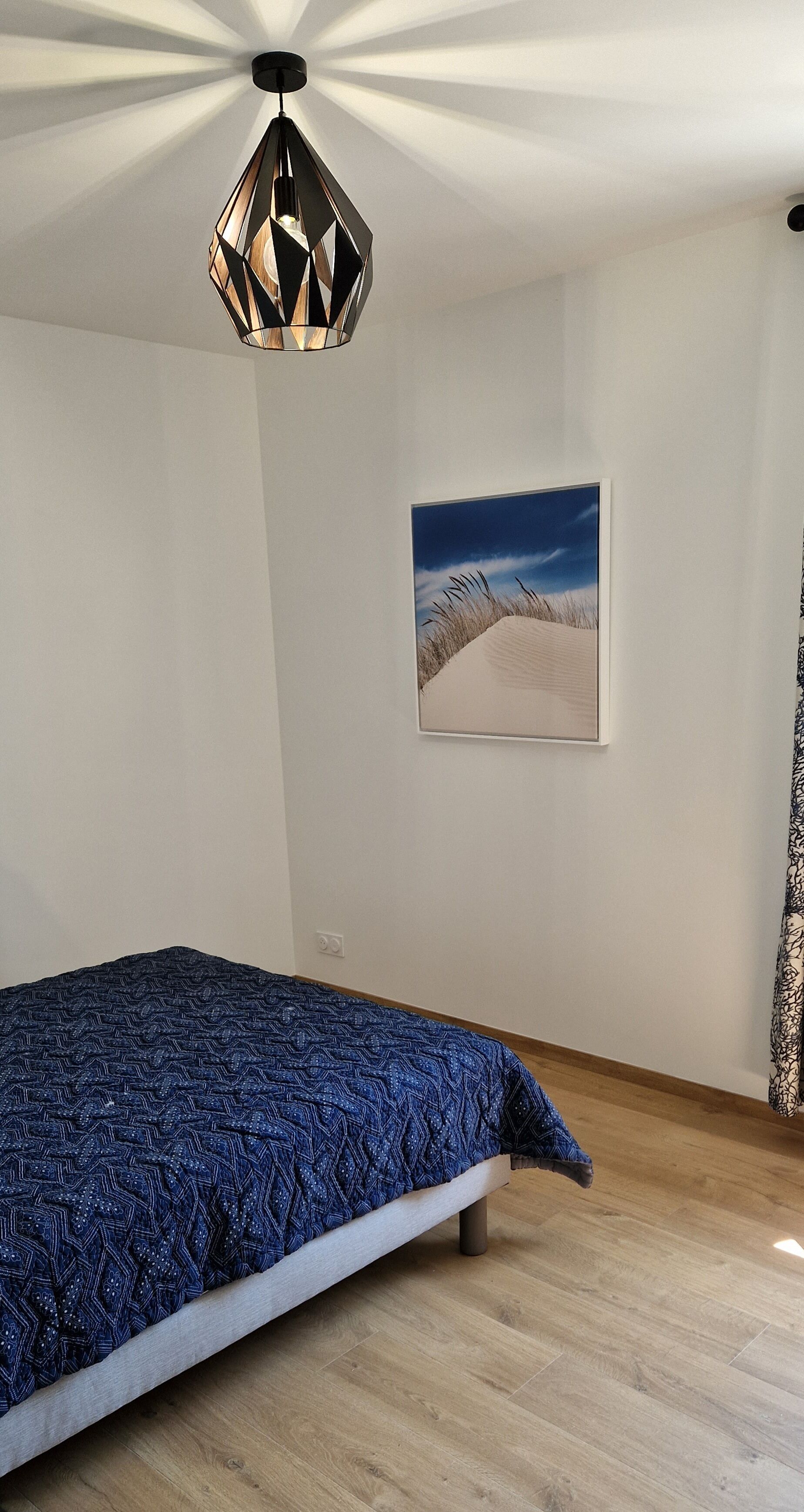 A bedroom with a bed, a geometric pendant light, a framed landscape painting, and a wooden floor.
