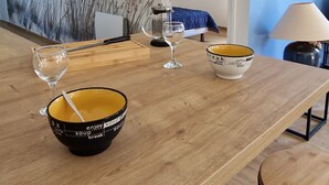 Un coin repas avec une table en bois, des bols et des verres. Un tableau représentant des roseaux est accroché au mur, et une lampe se trouve à droite.