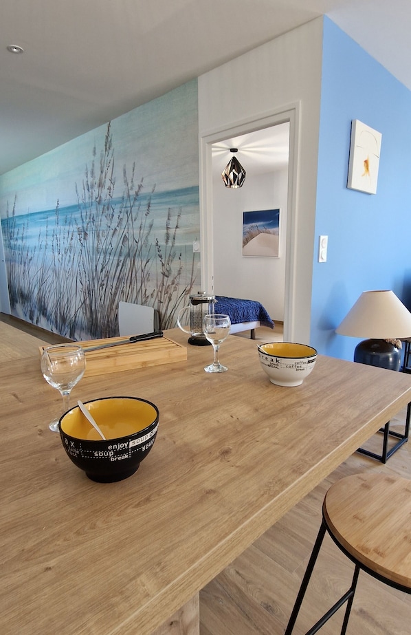 A dining area with a wooden table, bowls, and glasses. A painting of reeds is on the wall, and a lamp is on the right.