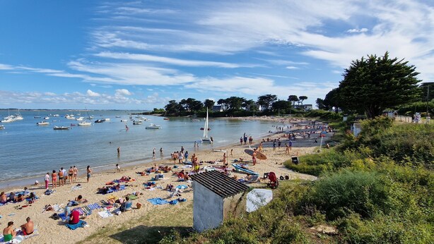 Une plage où l’on peut voir des gens se prélasser au soleil et nager, des bateaux sur l’eau, et une vue sur la mer et la côte lointaine.