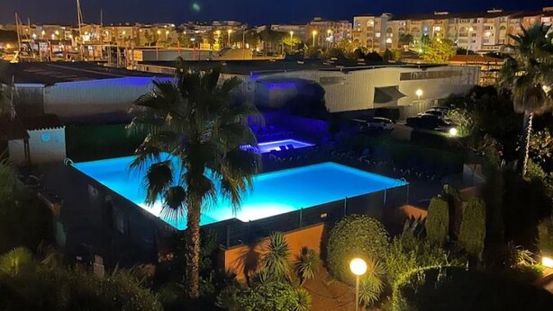 Ein Swimmingpool mit Wasserfall, umgeben von Palmen und einem gut beleuchteten Gebäudekomplex.