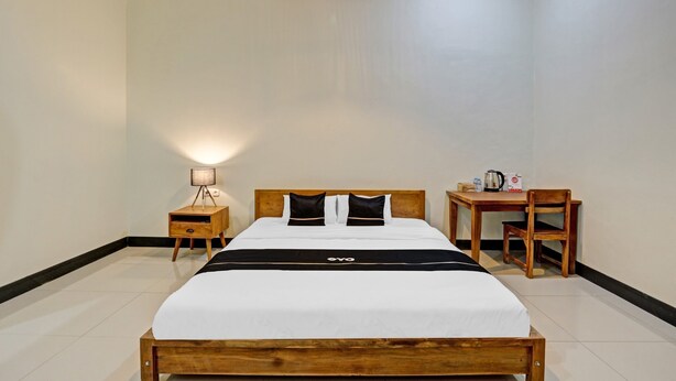 A hotel room with a wooden bed, a bedside table with a lamp, a small desk with a chair, and a hanging light fixture.