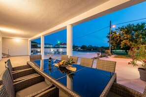 An outdoor dining area with a glass table and wicker chairs, overlooking a swimming pool and a landscaped garden.