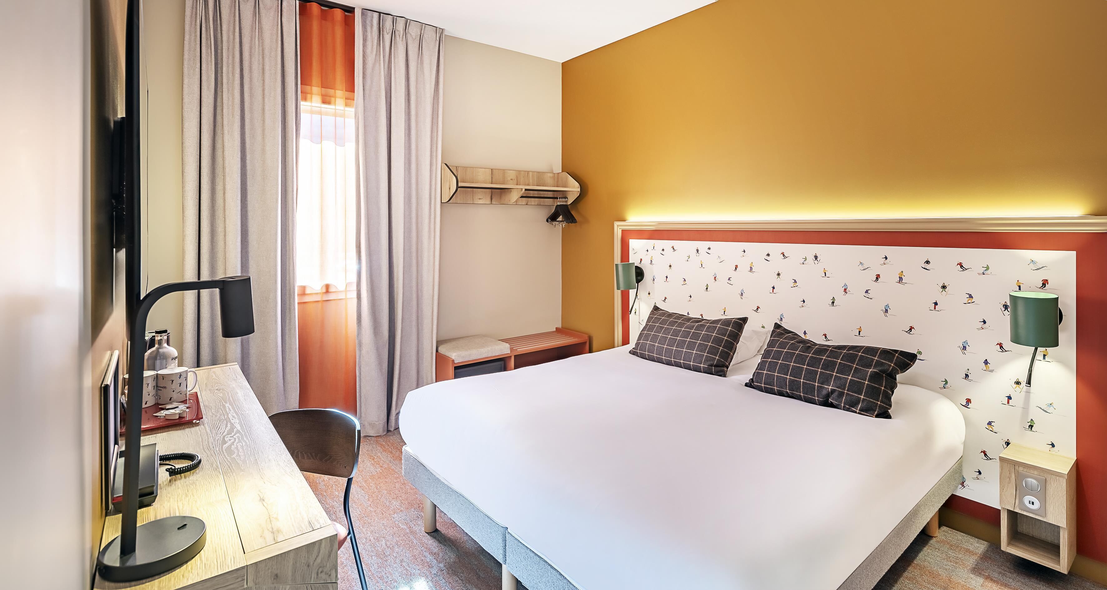 A hotel room with a bed, desk, chair, lamp, and a wall with a patterned wallpaper.