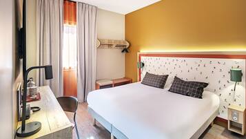 A hotel room with a bed, desk, chair, lamp, and a wall with a patterned wallpaper.