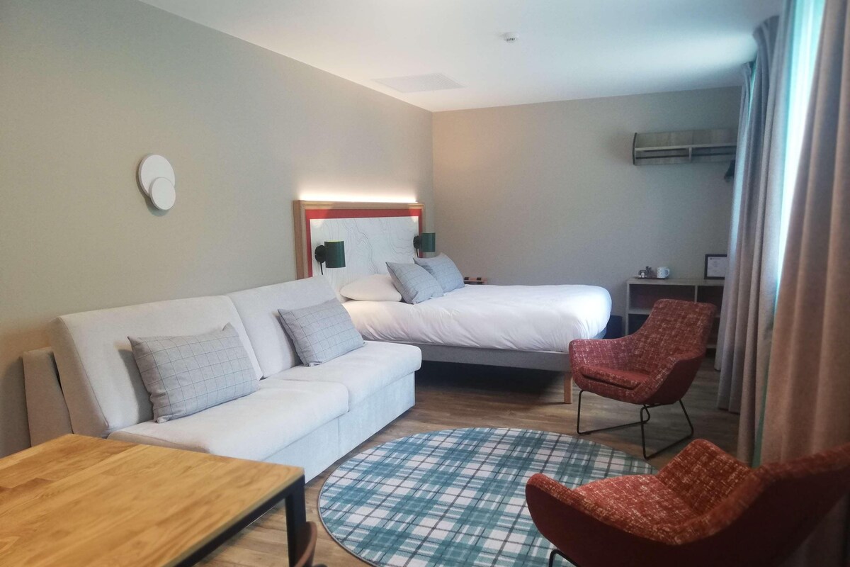 A modern hotel room with a white sofa, a bed, a red chair, and a wooden table.