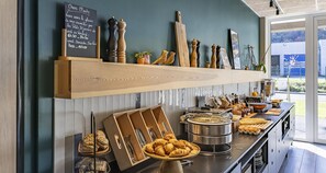 Una mesa de buffet con diversas opciones de comida y cubiertos, un menú en una pizarra y saleros y pimenteros decorativos en una estantería.