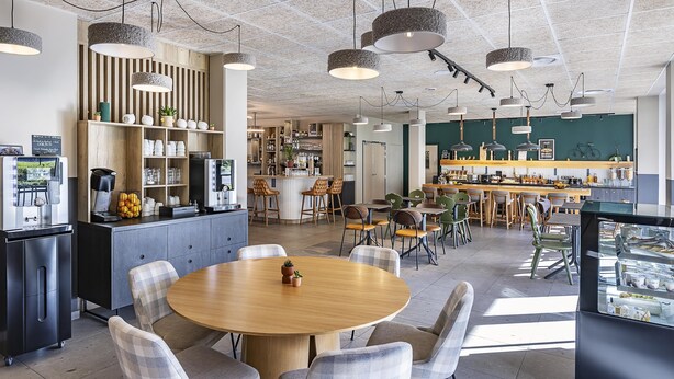 A modern cafe with a central round table, multiple seating options, and a variety of decor including plants and shelves with items.