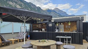 An outdoor seating area with a bar, wooden tables, and cushioned stools. - Best Western Coeur de Maurienne Hotel Restaurant & Spa (Saint-Jean-de-Maurienne)