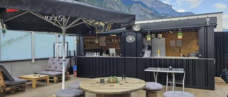 Un espace extérieur aménagé avec un bar, des tables en bois et des tabourets rembourrés.