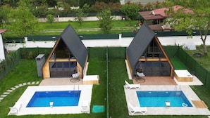 A symmetrical outdoor pool area with two separate pools, each with a lounge chair and a small deck. The pools are surrounded by well-maintained grass and a white fence. In the background, there are hills and a residential area with houses. - Sapanca Rose Garden Bungalov (Sapanca)