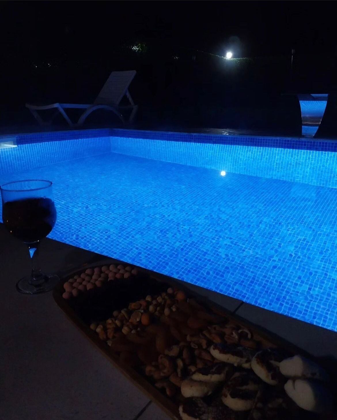 A swimming pool at night with blue lighting, a glass of red wine, and a tray of snacks.