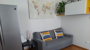 A compact living space with a grey sofa, two colorful cushions, a small table with a world map and a bottle of reed diffuser, a refrigerator, and a plant on a shelf. - Apartment in historic villa 10 minutes from the historic center (Firenze)