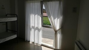 A room with a bunk bed, a window with white curtains, and a view of a road and greenery outside.