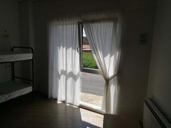 Una habitación con literas, una ventana con cortinas blancas y vistas a una carretera y vegetación.