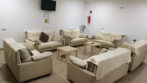 A room with beige sofas arranged in a semi-circle, a flat-screen TV mounted on the wall, a fire extinguisher, and a small table with a fan.