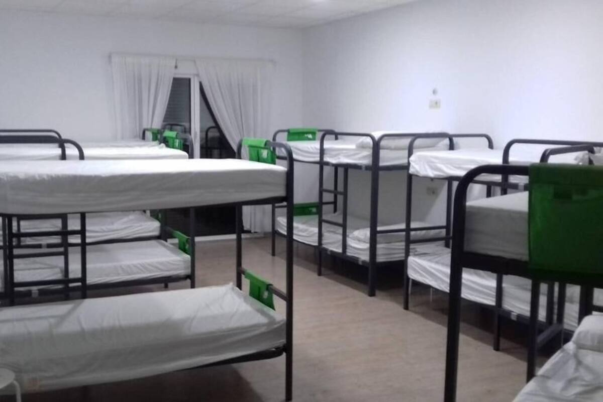 A dormitory room with bunk beds, a window with curtains, and a small table with a chair.