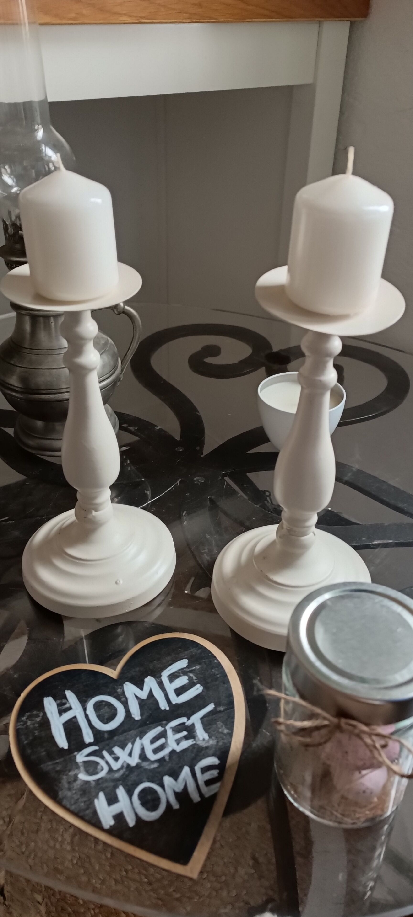 A kitchen counter with two white candle holders, a heart-shaped sign with 'HOME SWEET HOME,' and a glass jar with a metal lid.